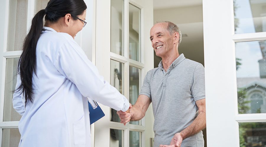 Female doctor with clipboard shaking hand of senior man during home visit while entering house